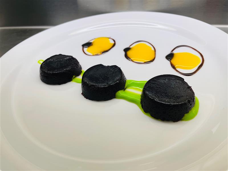 Trois bouchées rondes de bleuets à manger-main, de couleur bleu très foncé, présentées sur une grande assiette blanche avec une sauce verte et trois touches de coulis jaune entourées d’un filet foncé.