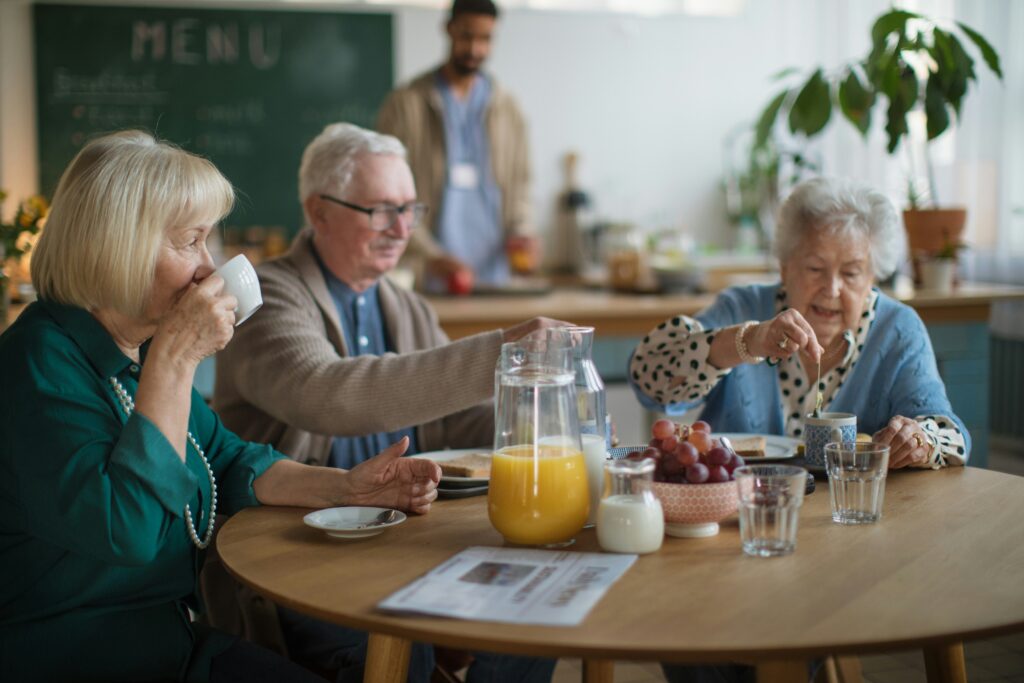 Trois personnes âgées prennent un déjeuner autour d’une table ronde dans une salle à manger communautaire lumineuse. Sur la table se trouvent un pichet de jus d’orange, du lait, des raisins, des verres d’eau et des assiettes de déjeuner.