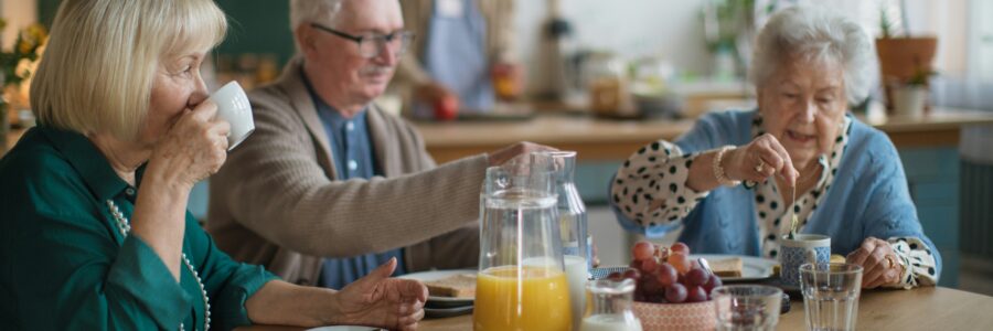 Trois personnes âgées prennent un déjeuner autour d’une table ronde dans une salle à manger communautaire lumineuse. Sur la table se trouvent un pichet de jus d’orange, du lait, des raisins, des verres d’eau et des assiettes de déjeuner.