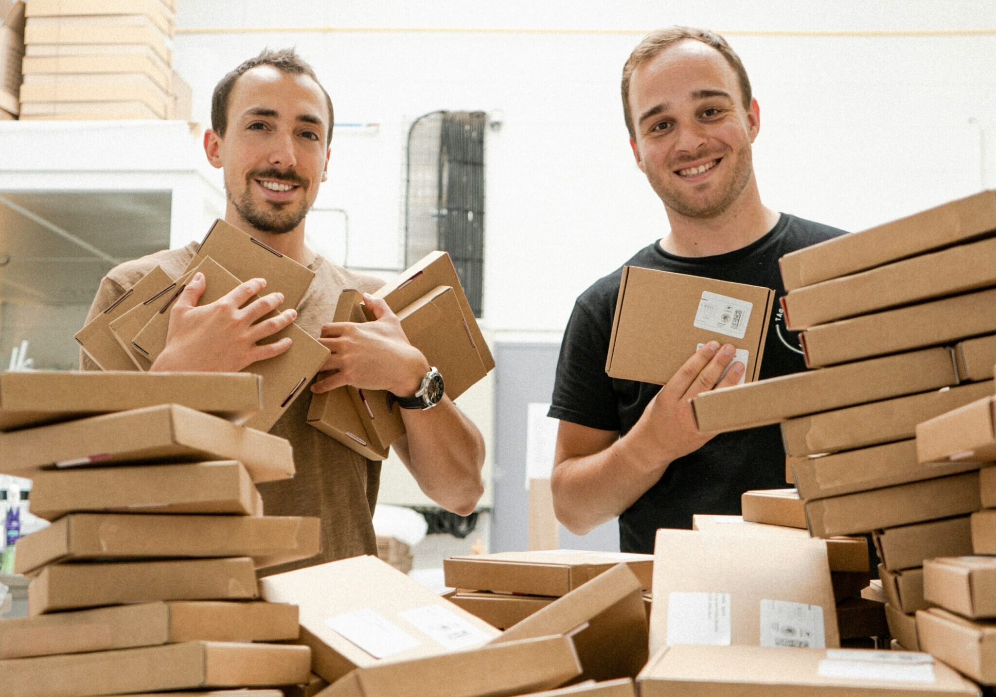 Deux entrepreneurs souriants tiennent des boîtes d'expédition, entourés de piles de colis dans l'entrepôt de leur PME